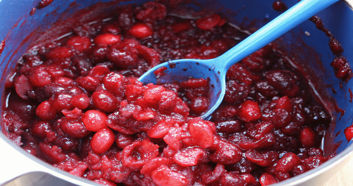 Close-up view of a blue pot filled with homemade cranberry sauce. The sauce has a thick, glossy texture with whole cranberries and chunks visible. A blue spoon is scooping out a portion of the sauce.