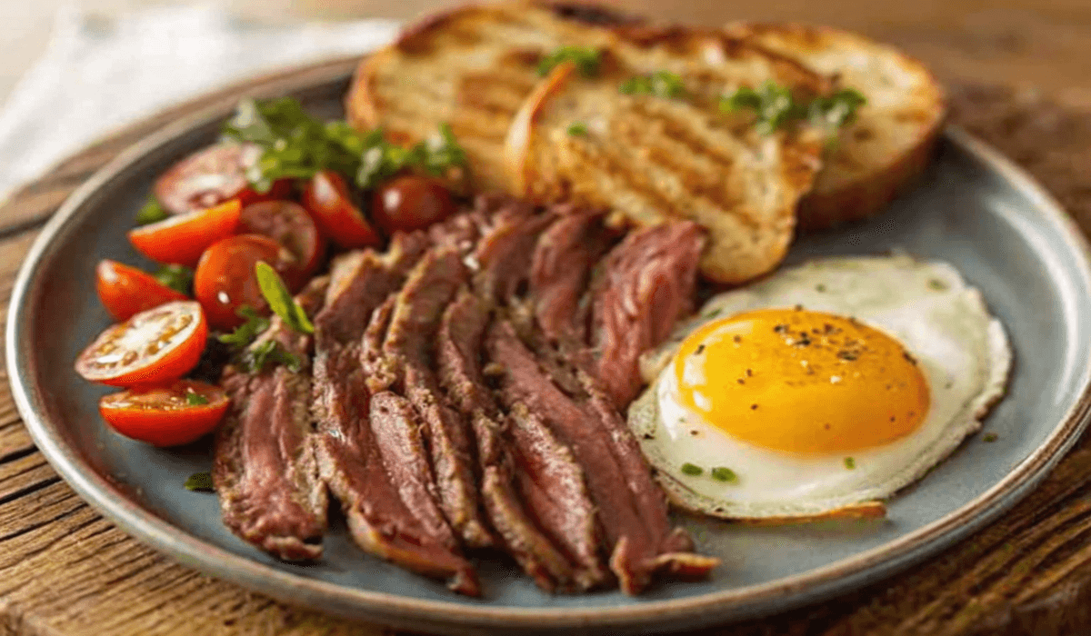 A breakfast plate with sliced steak, a sunny-side-up egg, a salad of cherry tomatoes and greens, and two slices of grilled toast on a rustic wooden table.